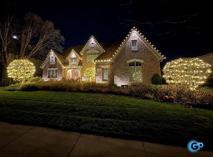 Warm white roofline lighting with wrapped shrubs on a brick two-story home at night