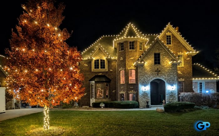 A fully wrapped front-yard tree beside a home outlined in warm white roofline lighting